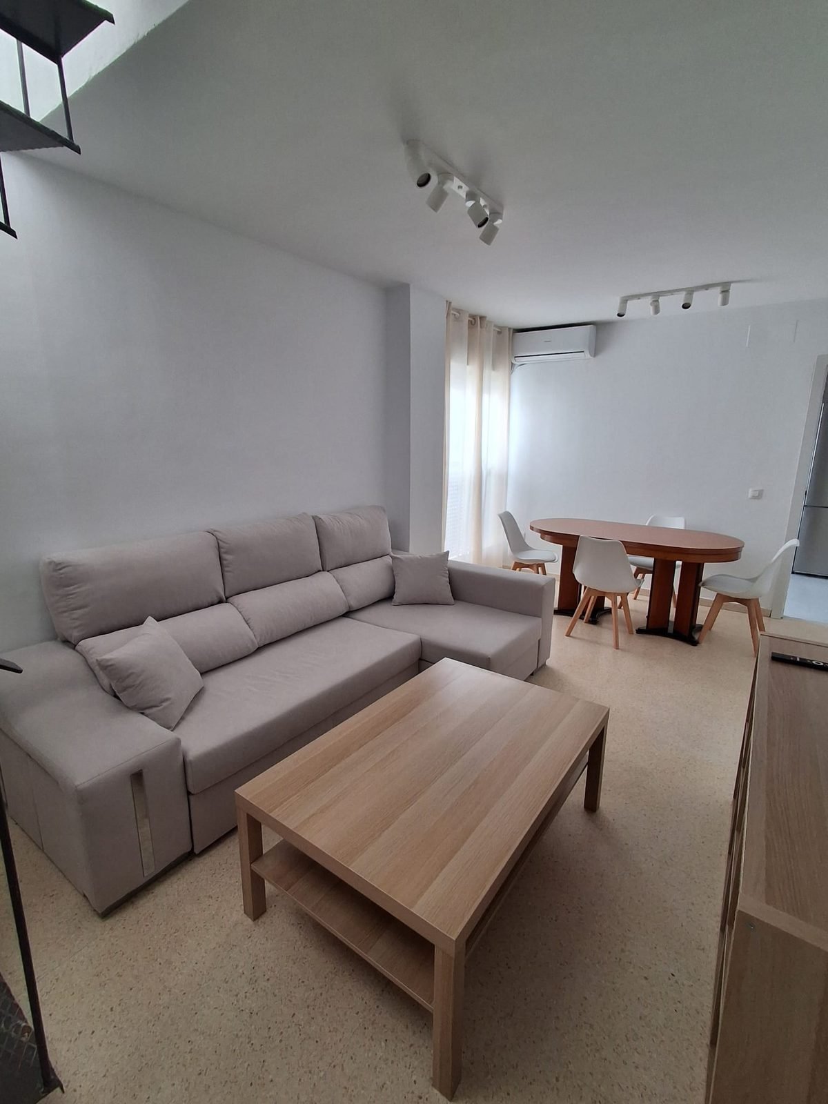 Salón con sofá claro, mesa de centro de madera y comedor junto a la ventana